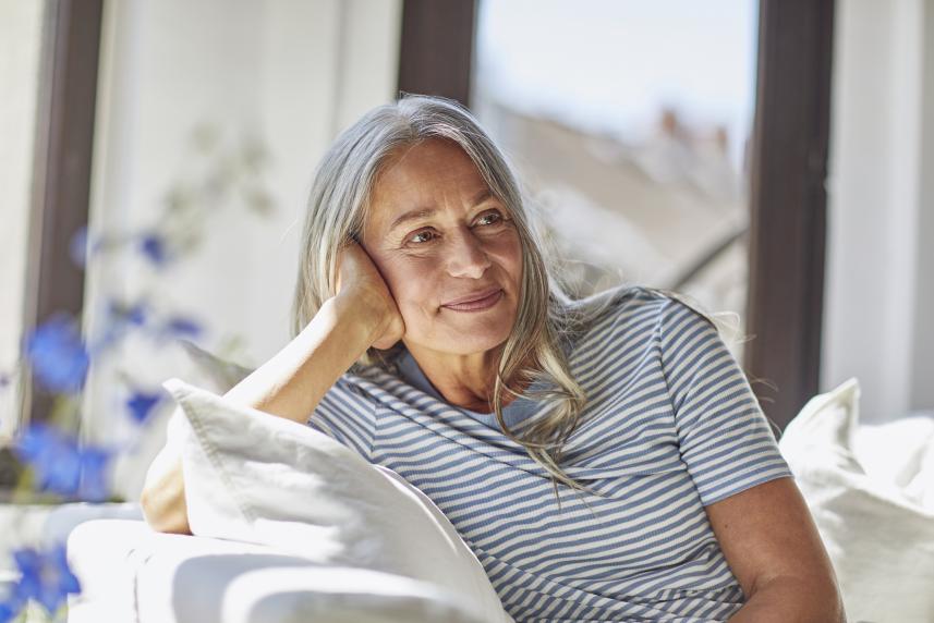 A woman with long, grey hair smiles slightly as she sits on a couch, her hand on the side of her head and her elbow resting on the armrest. She is in a room full of natural light and white furniture.