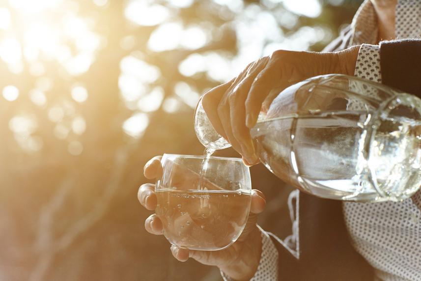 Hands of senior woman pouring glass of water by window - stock photo