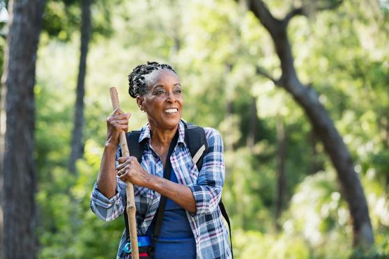 A senior African-American woman in her 70s hiking in a park smiling and leaning on a walking stick, standing with trees and lush foliage in the background.