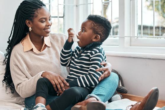 A child in a striped sweater sits on his mother's lap as he uses an inhaler. She is smiling at him. Both mother and son have medium brown skin and dark black hair.