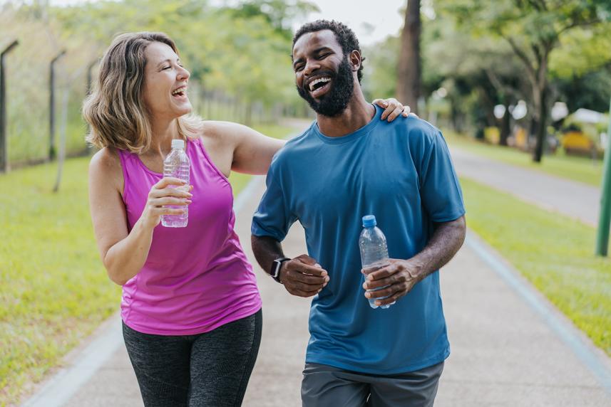 A woman and man laugh while walking on a trail together. They're wearing athletic clothing and holding plastic water bottles. Her hand is on his shoulder.