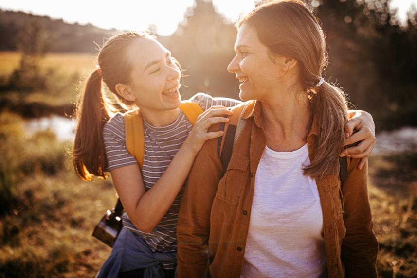 A woman and her daughter smile at each other as they walk through nature during golden hour.