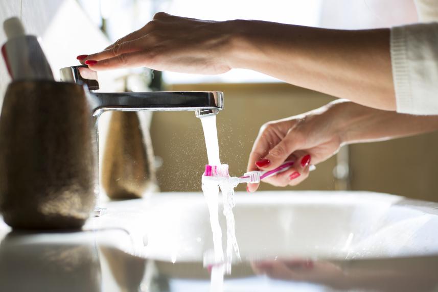 A person with red-painted fingernails holds a pink toothbrush under a running sink faucet.