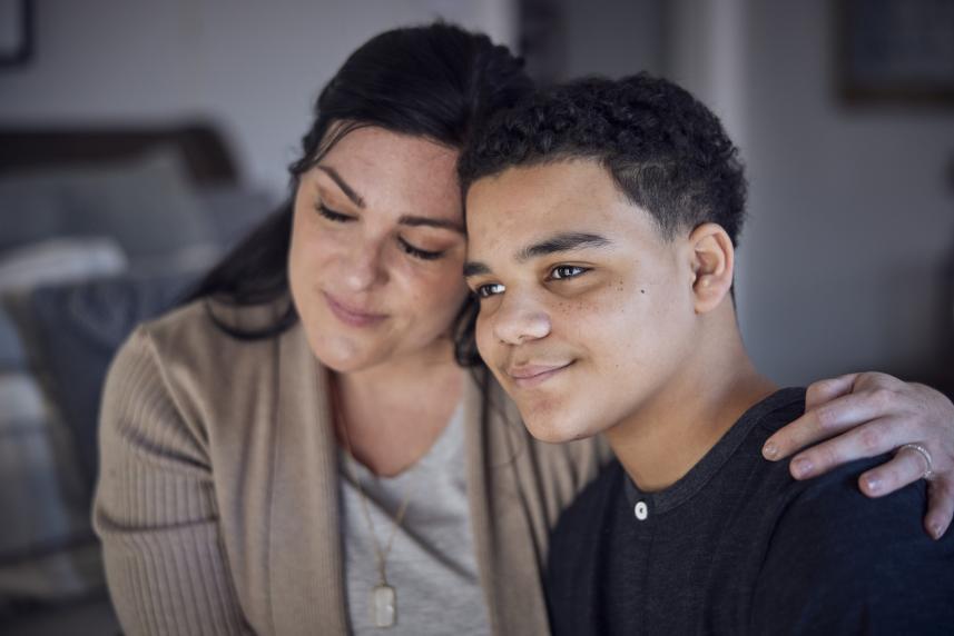 A mother sits beside her son with her arm around his back, her hand resting on his shoulder. The scene feels content and peaceful. They both have slight smiles and look very relaxed.