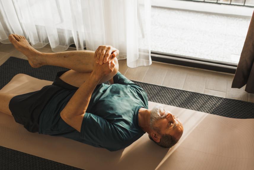 A man lays face-up on a yoga mat and pulls his right knee to his chest to stretch his lower back.