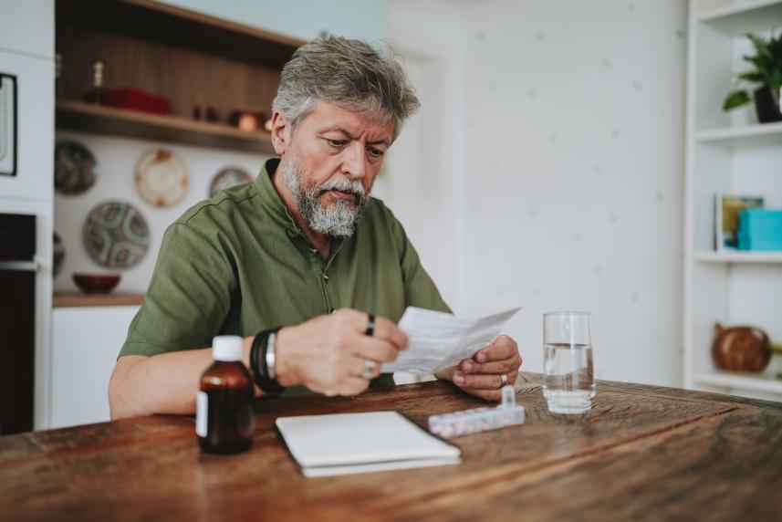 An older man reads medical paperwork at his kitchen table.