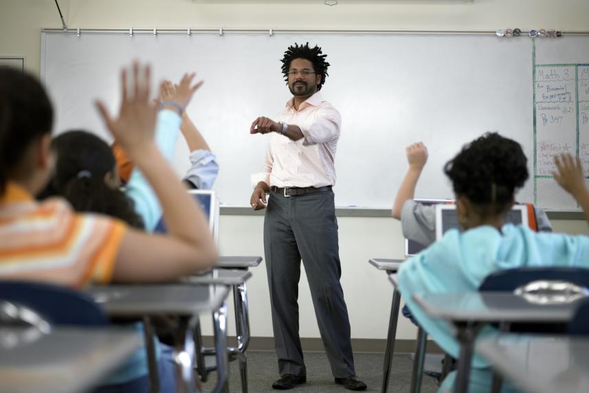 A teacher stands at the front of his middle school classroom as his students sit before him with their hands raised.