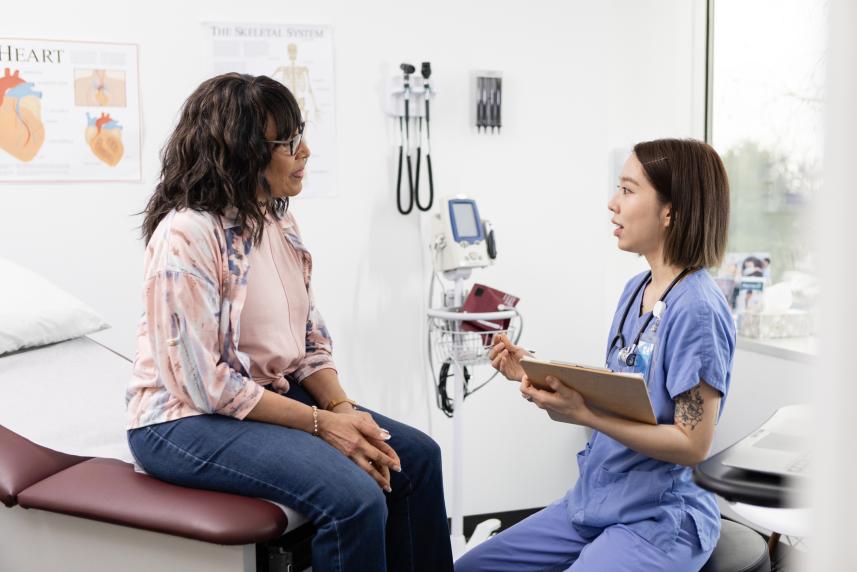 A woman sits on an exam chair at a doctor's office while a doctor in blue scrubs takes notes while they talk.