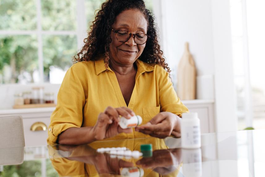 A middle-aged Black woman in a yellow blouse sits at a table and smiles gently as she sorts through her medications.