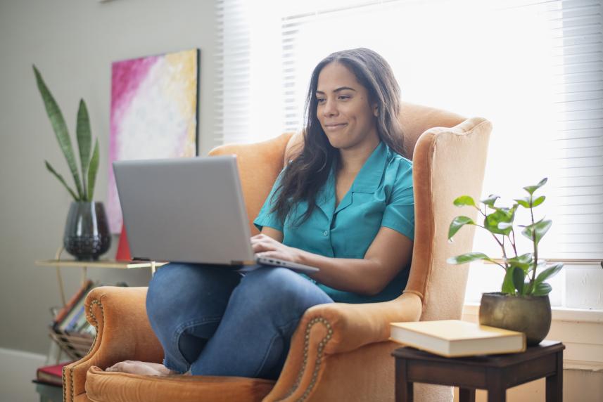 A woman sits in an armchair with a laptop resting on her knees.