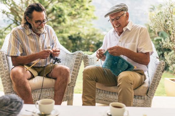 Two older men sit outside and knit together.