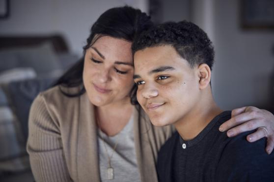 A mother sits beside her son with her arm around his back, her hand resting on his shoulder. The scene feels content and peaceful. They both have slight smiles and look very relaxed.