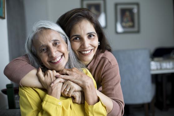 A daughter hugs her mother from behind, and her mother folds her own hands over her daughter's. Both are smiling at the camera.