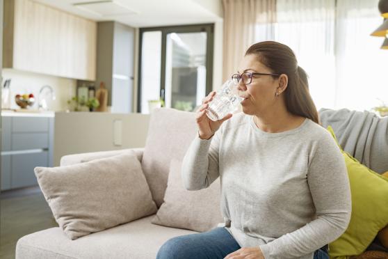 A brunette woman sits on her sofa and sips a glass of water.