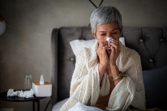 A middle-aged woman sits in bed blowing her nose, wrapped in a blanket, with crumpled up tissues on her night stand.