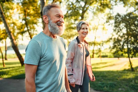 An older couple smile while walking down a sunlit sidewalk near a body of water.