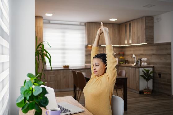 A woman in a yellow blouse sits at her home-office desk with both arms raised in a stretch above her head.