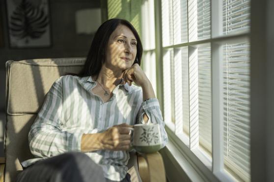 A woman looks pensively out the window while sitting in an armchair and holding a mug.