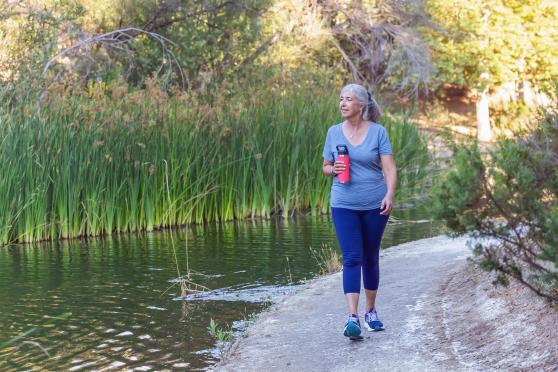 A woman walks on a path along a river while holding a pink water bottle.