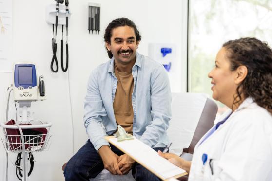 A man sits on an exam table at the doctor's office while talking to his provider during his annual checkup.