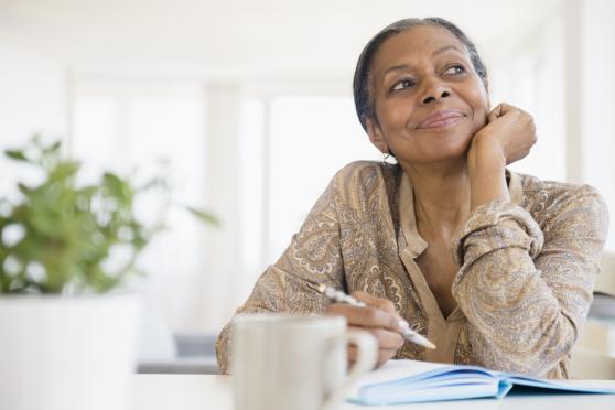 A woman sits pensively at her kitchen table, pen in hand, with a notebook and a coffee mug before her.