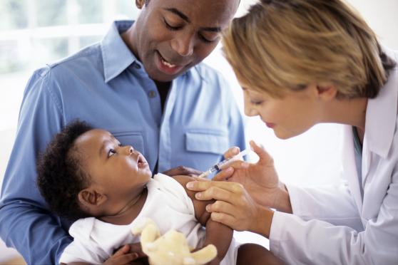 A man holds his young baby while a doctor administers a vaccination to the baby.