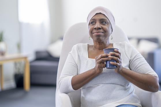 A woman sips a cup of tea while she undergoes cancer treatment. She wears a scarf around her head.