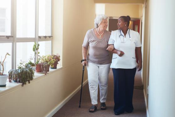 Photo: Senior woman walking with cane in hallway with assistance from healthcare worker.