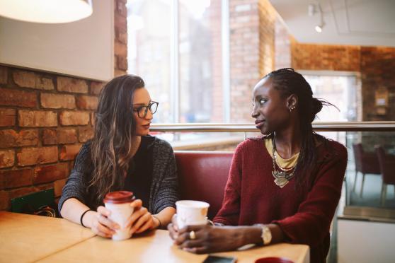 Two women sit at a table together and talk over coffee.