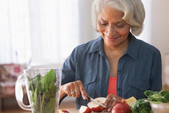 A woman with short white hair slices an apple to toss into the blender on the counter beside her. The blender is already filled with leafy greens.